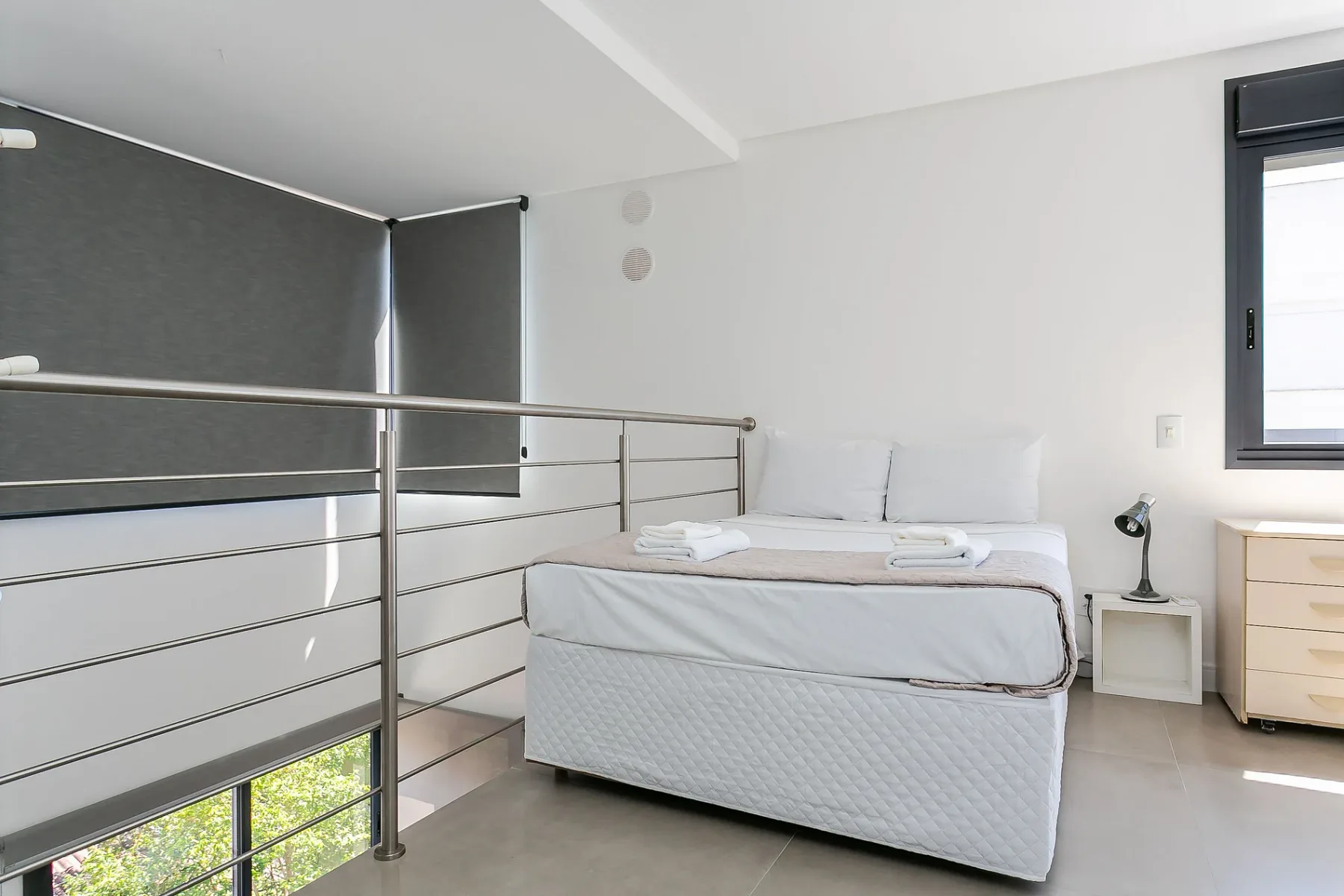 A bright bedroom with a neatly made bed beside a metal railing.