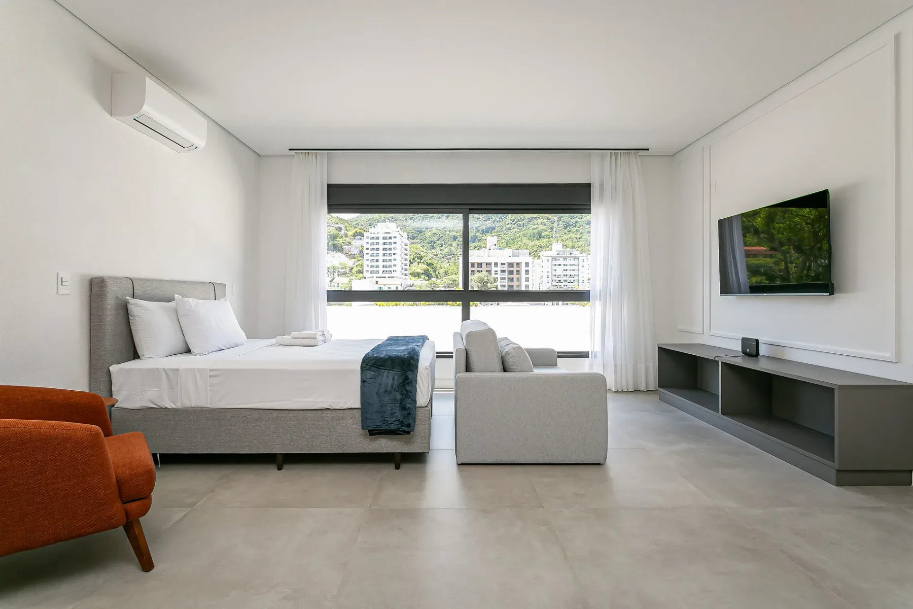 A bright bedroom with a bed, armchair, and large window overlooking city buildings.