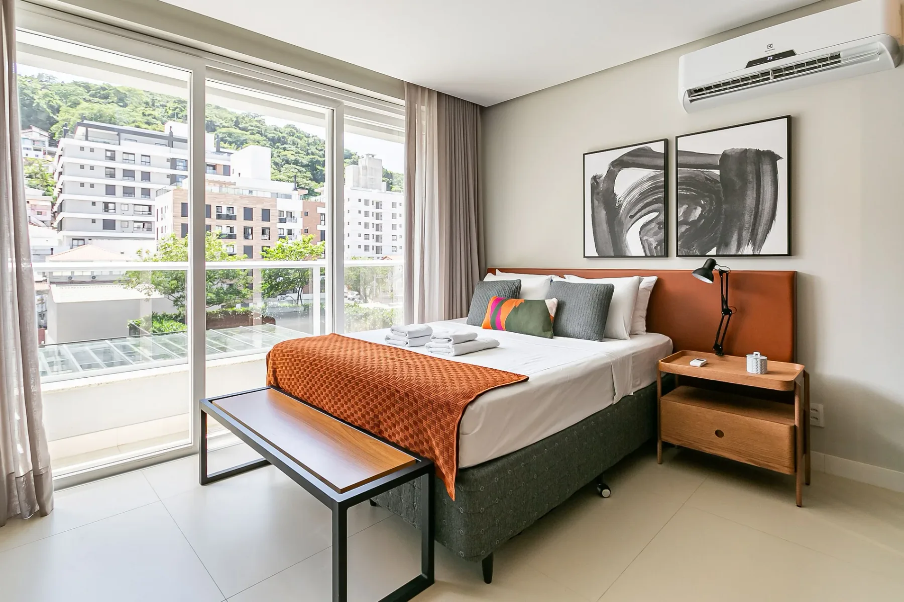A bright bedroom with a bed beside large windows overlooking nearby buildings.