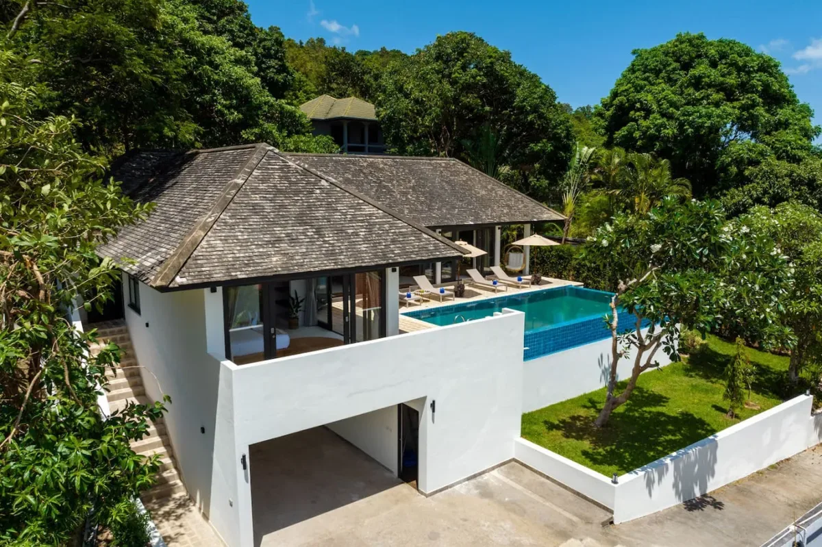 A white villa with a tiled roof overlooks a swimming pool surrounded by greenery.