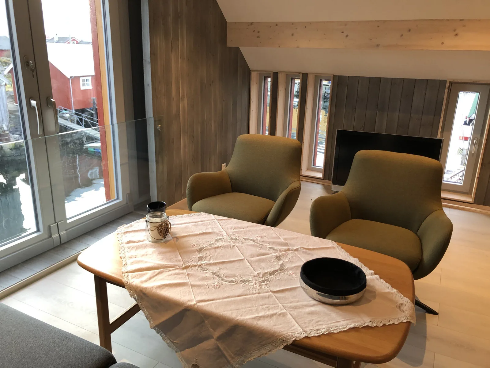Una zona de estar revestida de madera tiene dos sillones alrededor de una mesa junto a grandes ventanas.