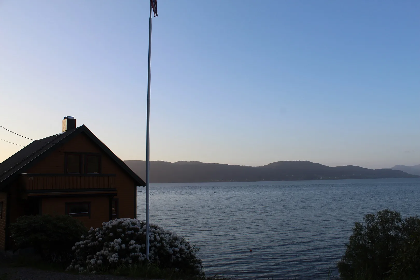 Escena junto a un lago, con una casa pequeña en primer plano y colinas al otro lado del agua.