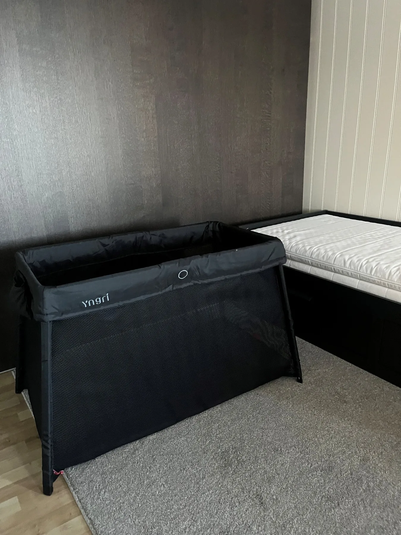 Dormitorio con un corralito negro de malla junto a una cama y una alfombra gris.