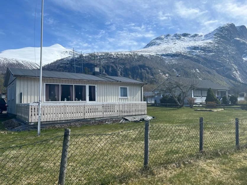 Una casa pequeña con patio cercado se encuentra bajo montañas nevadas.