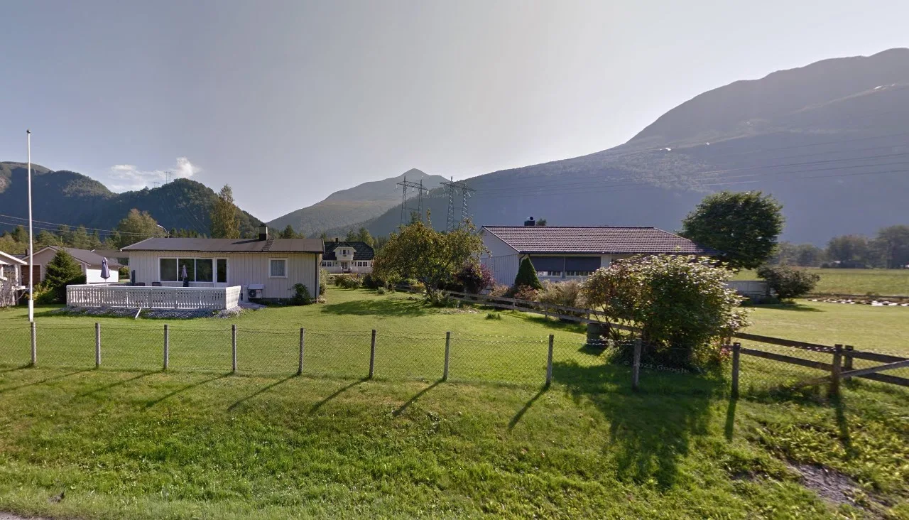 Una propiedad rural se encuentra junto a un campo de hierba cercado, con montañas al fondo.