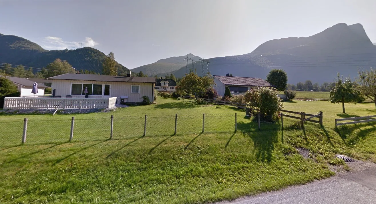 Una casa rural y edificios cercanos junto a un campo verde cercado, con montañas al fondo.