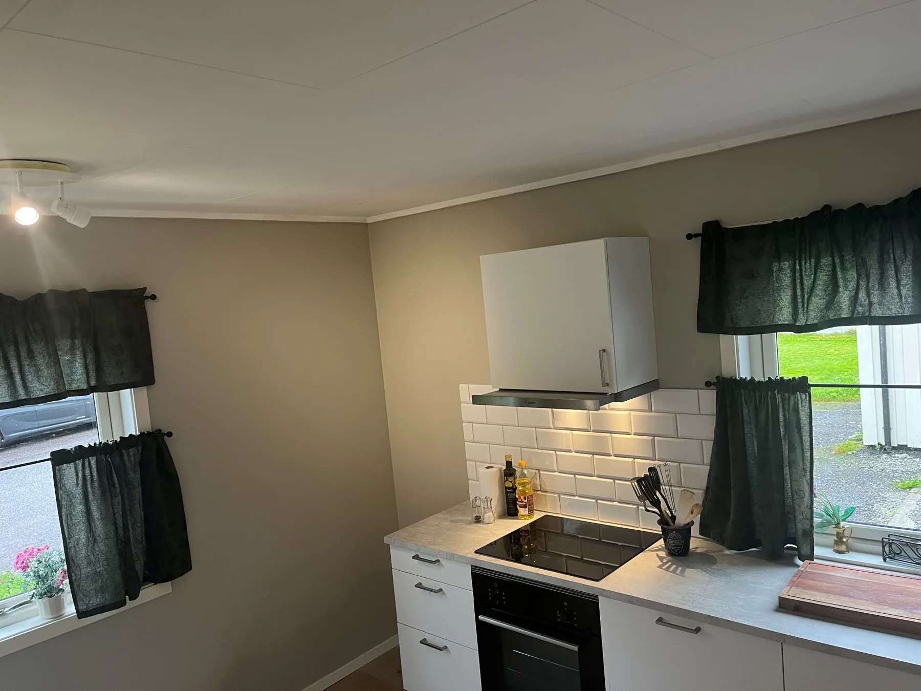 Una cocina compacta con gabinetes blancos, un salpicadero de azulejos y ventanas con cortinas oscuras.