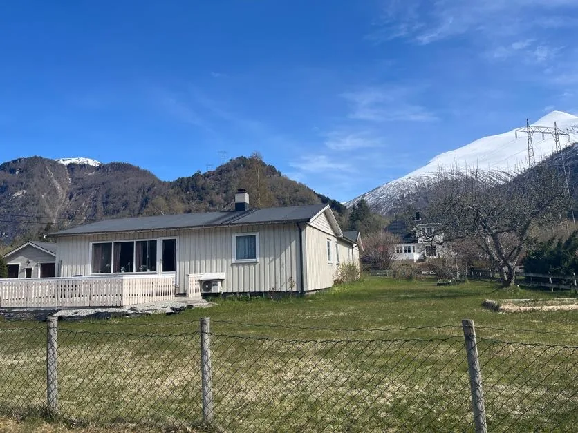 Una casa de color claro está detrás de un patio cercado con césped y montañas al fondo.
