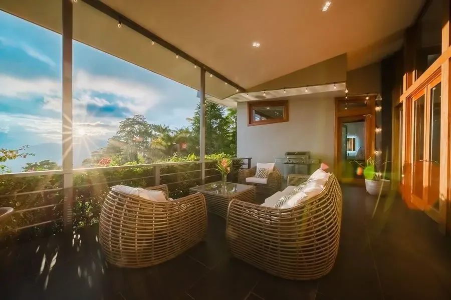 Una zona de estar exterior cubierta con asientos tejidos junto a grandes ventanales.