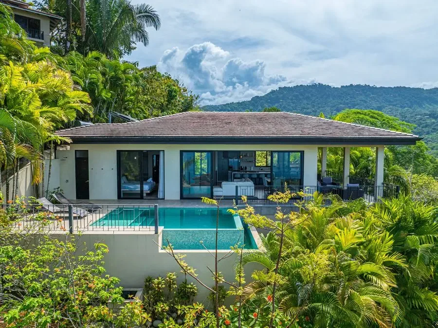 A modern house opens onto a swimming pool, surrounded by lush tropical greenery.