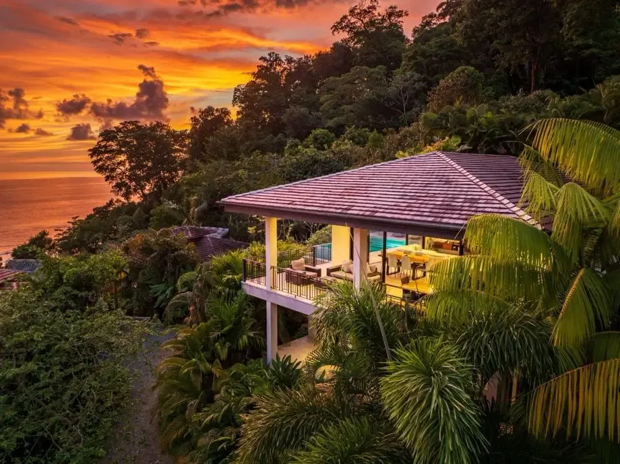 A hillside villa overlooks the ocean at sunset, surrounded by dense tropical greenery.