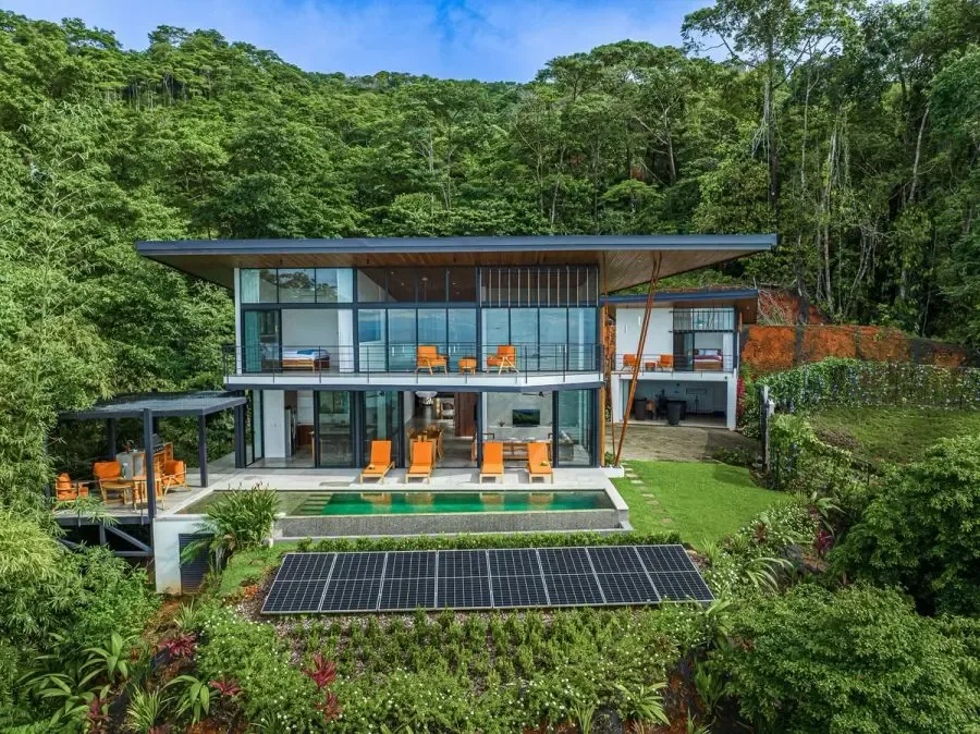 A modern glass-fronted house sits amid dense green forest, with a lawn and solar panels.