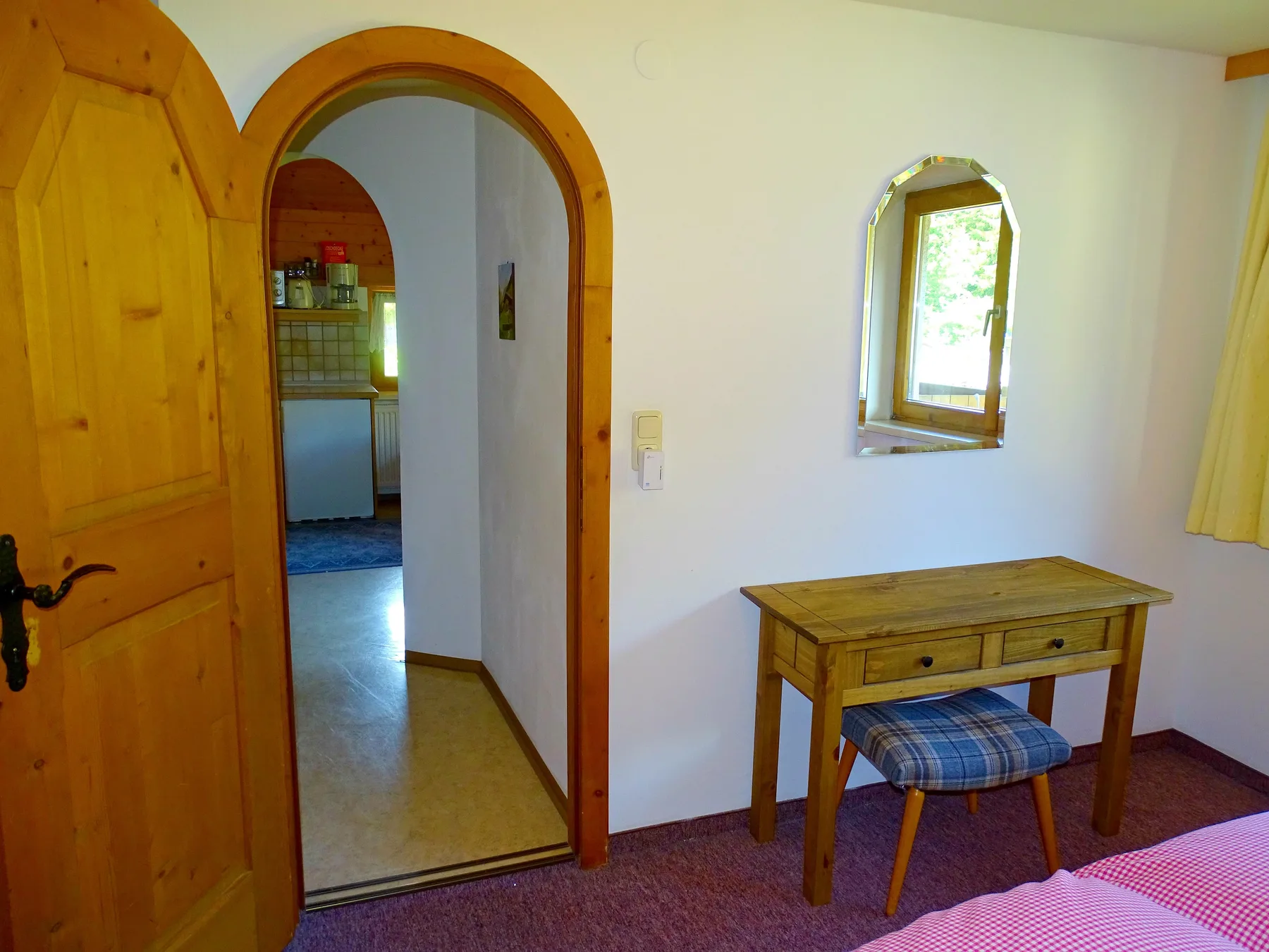 A bedroom with a wooden desk beneath a small window and an arched doorway.