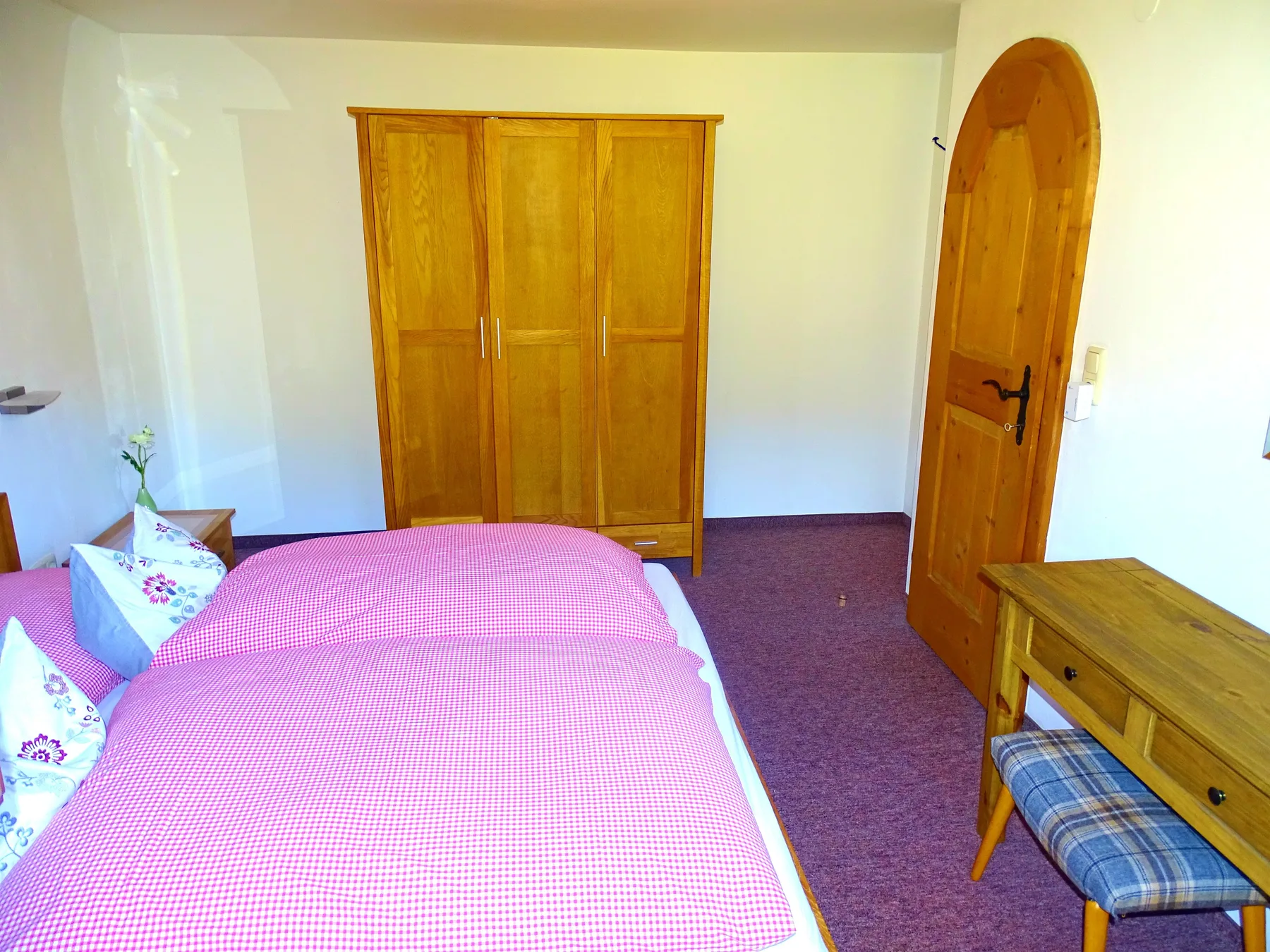 A bedroom with a pink bed, wooden wardrobe, and open wooden door.
