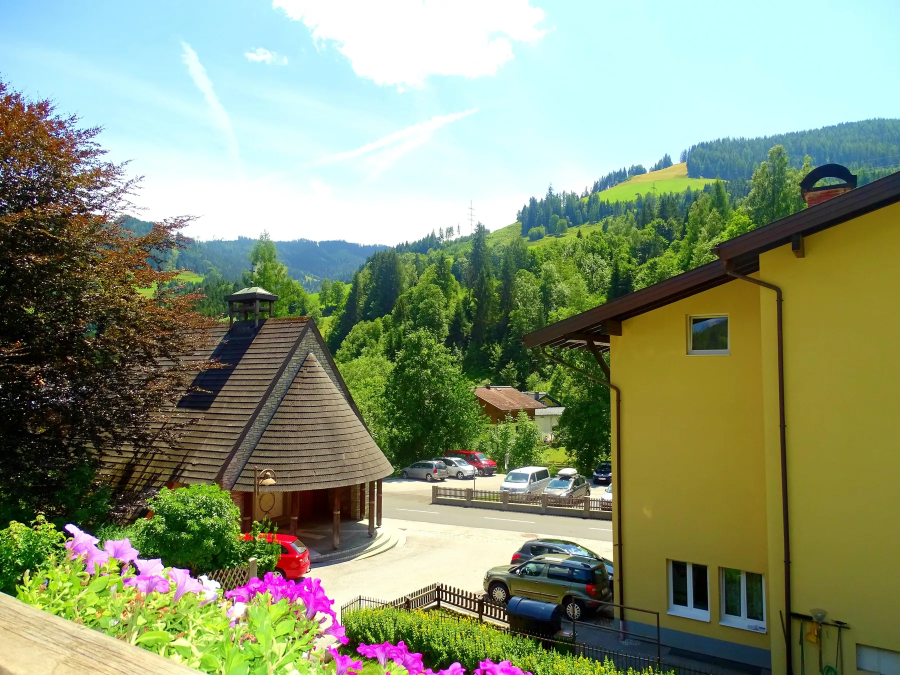 A roadside scene with colorful flowers, parked cars, and forested hills.