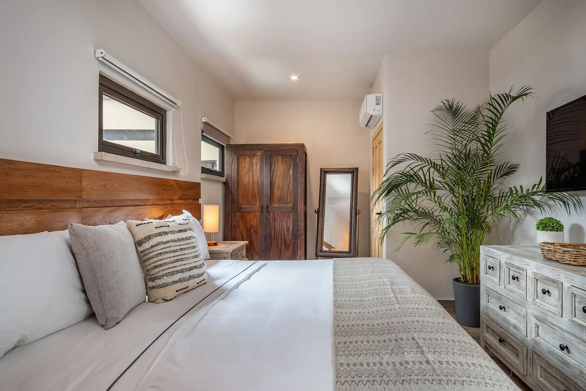 Un dormitorio luminoso con una cama bien tendida y una planta alta de hojas.
