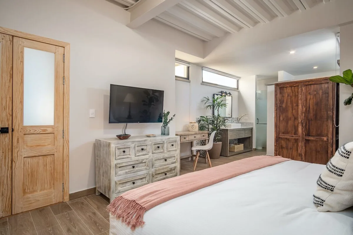 A bright bedroom with a bed, wooden dresser, and a desk by the windows.