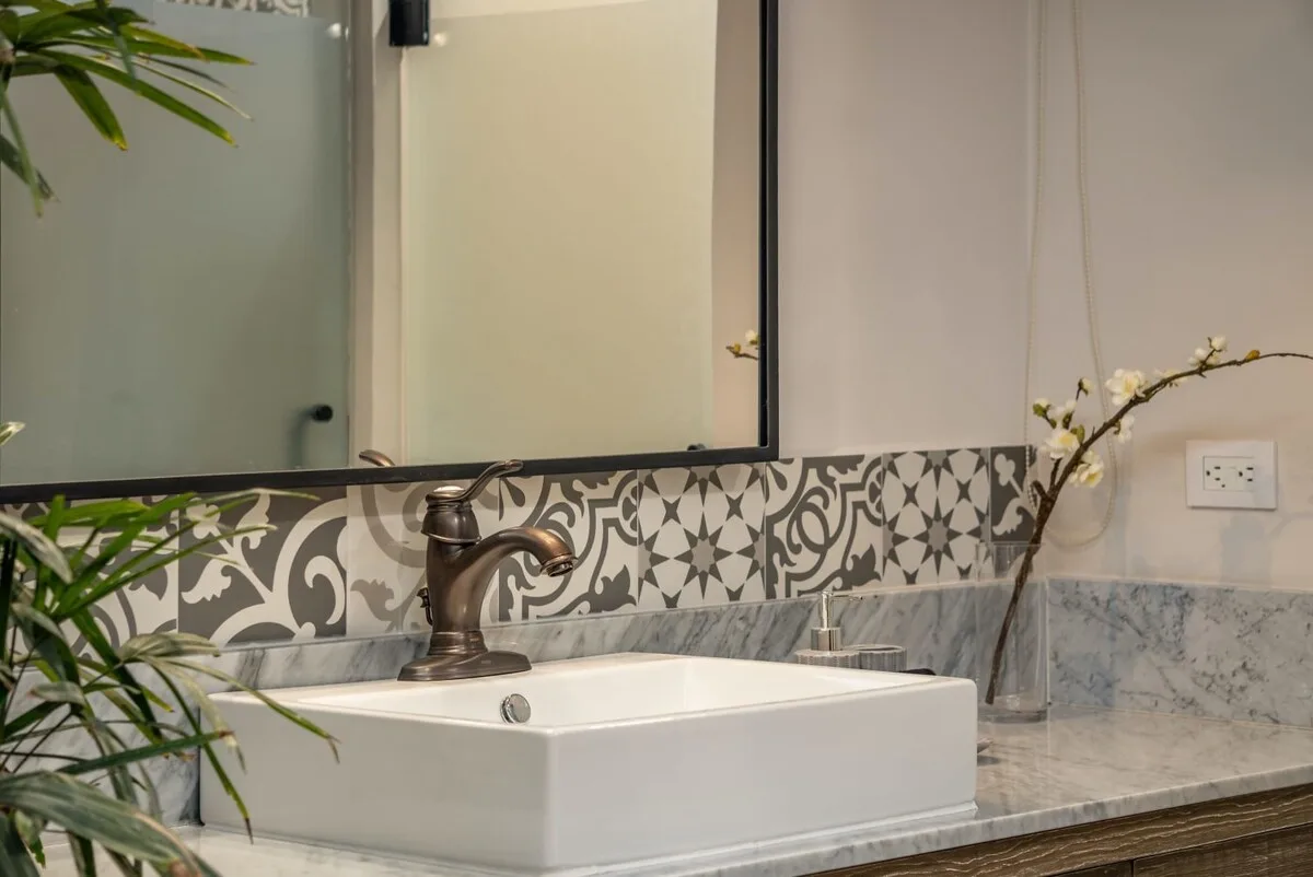 A bathroom vanity with a white basin, bronze faucet, and patterned backsplash.