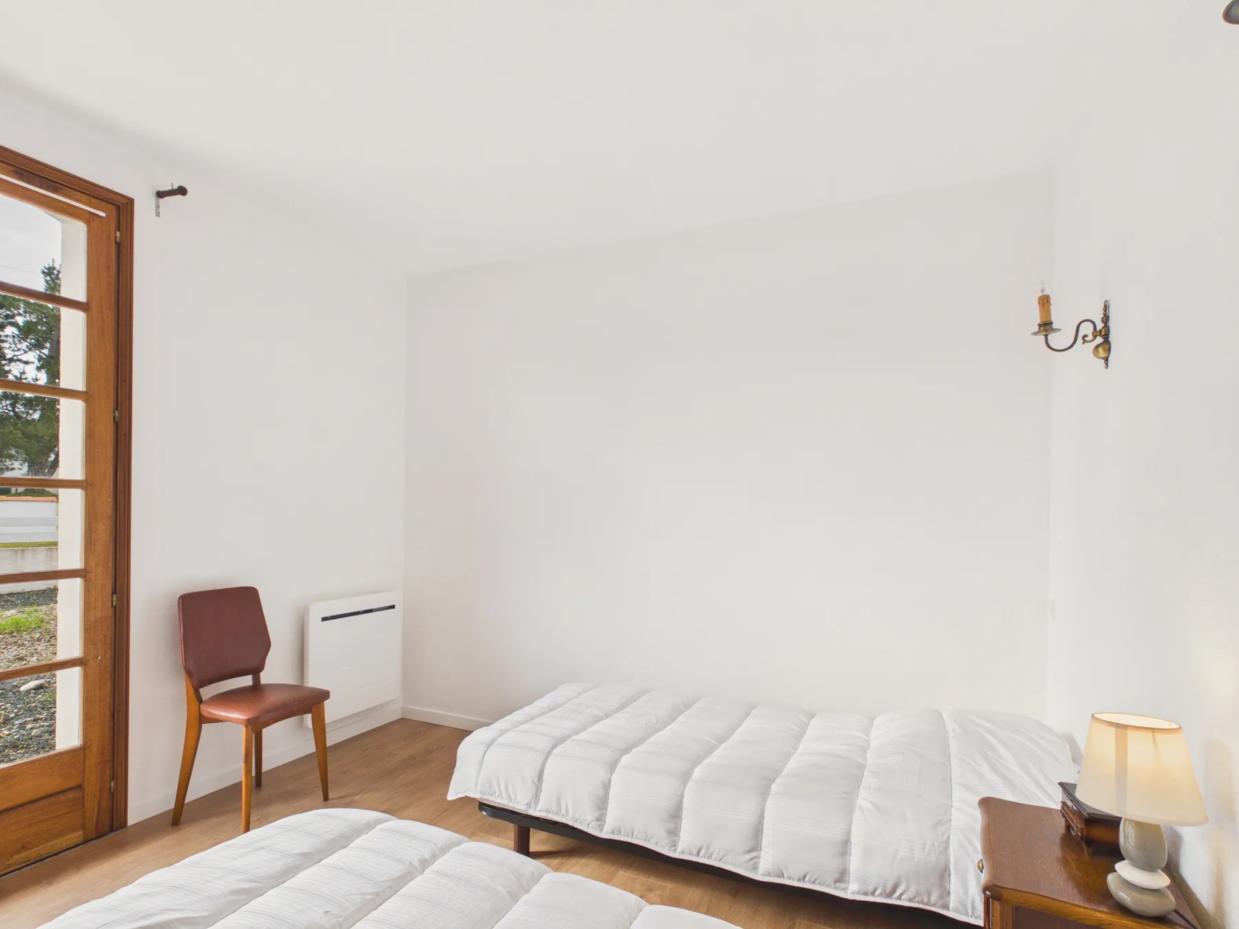 A bright bedroom with two beds, a chair, and a tall window.