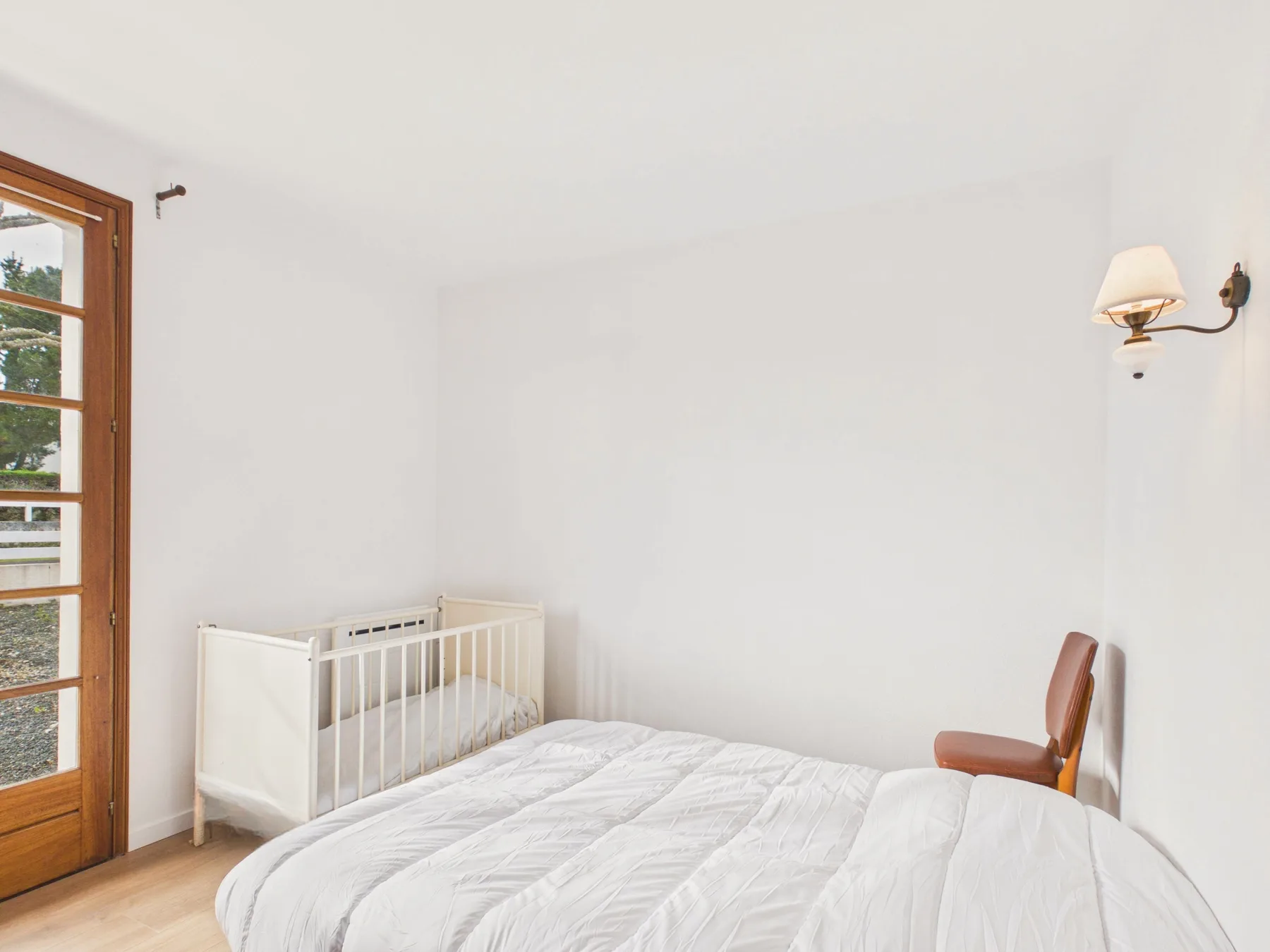 A bedroom with a white bed and a small crib beside a glass-paneled door.