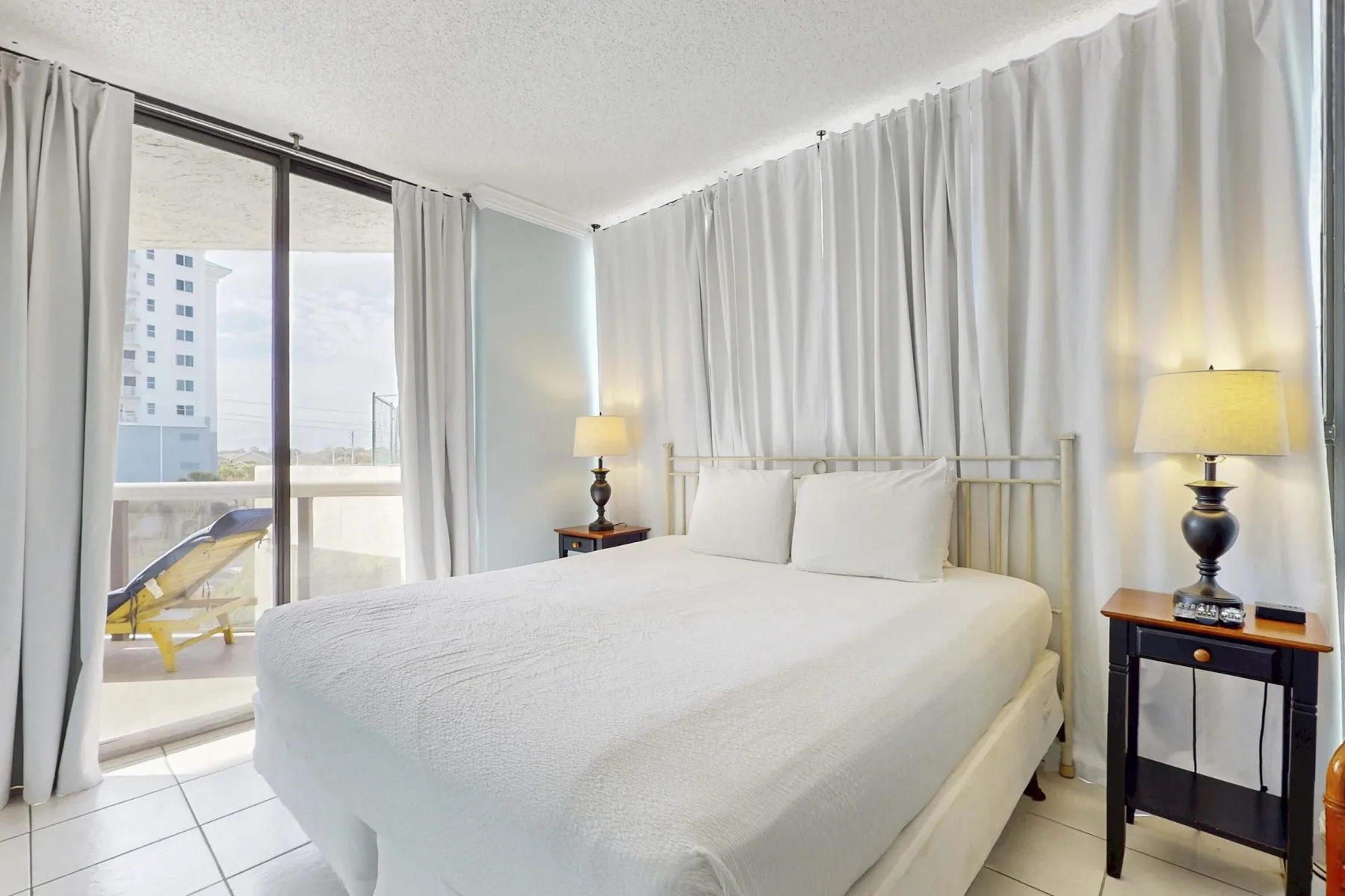 A bright bedroom with a neatly made bed beside a large curtained window.