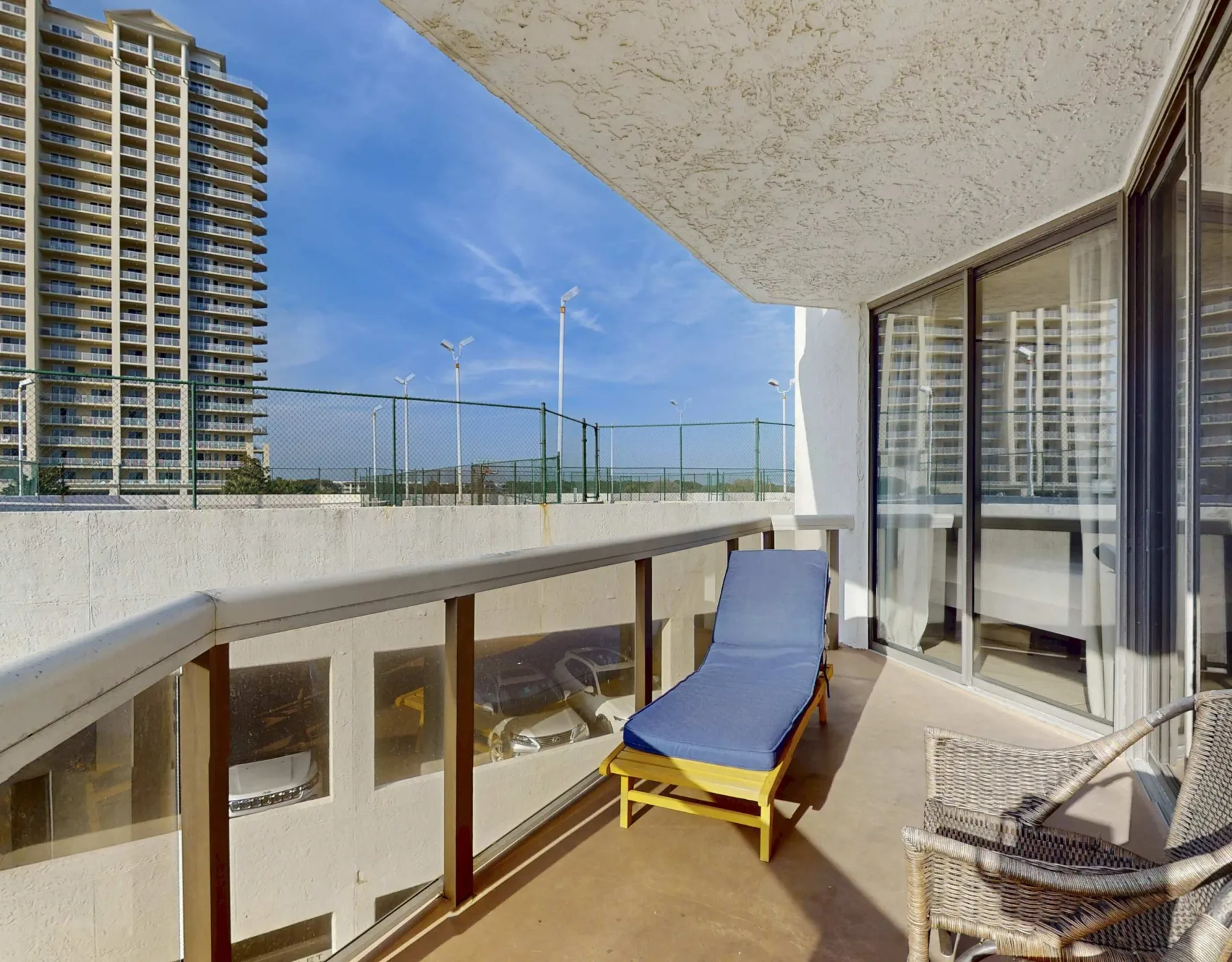 A sunny balcony with a blue lounge chair and glass doors overlooking nearby buildings.