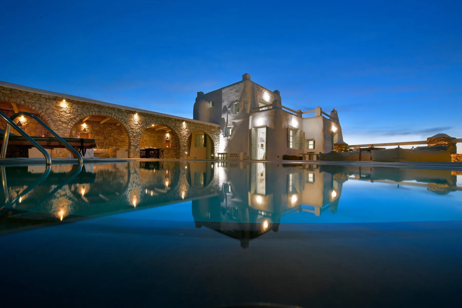 Un edificio iluminado y un muro con arcos se reflejan en una piscina al atardecer.