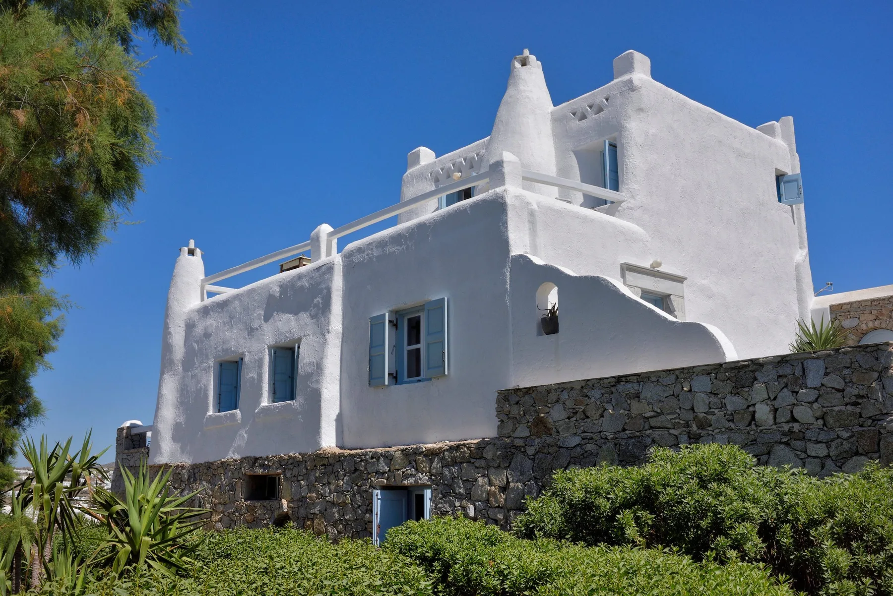 Un edificio blanco de estuco se alza sobre un muro bajo de piedra bajo un cielo azul despejado.