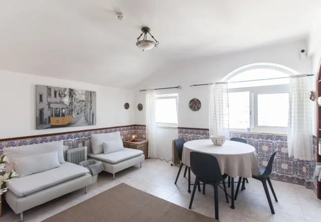 A bright sitting and dining area with light seating, a round table, and large windows.