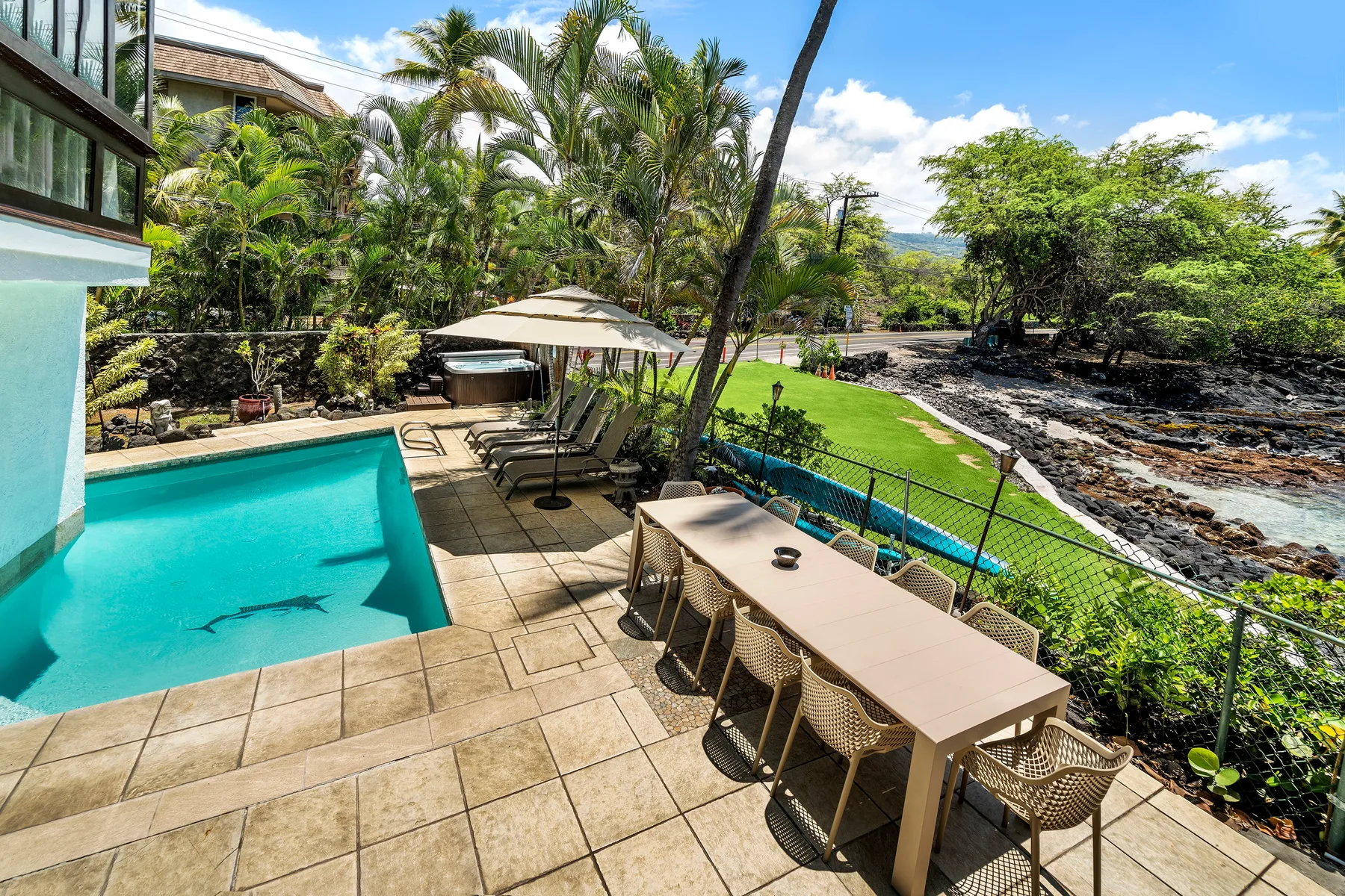 Una piscina exterior soleada y un patio con asientos junto a un césped verde.