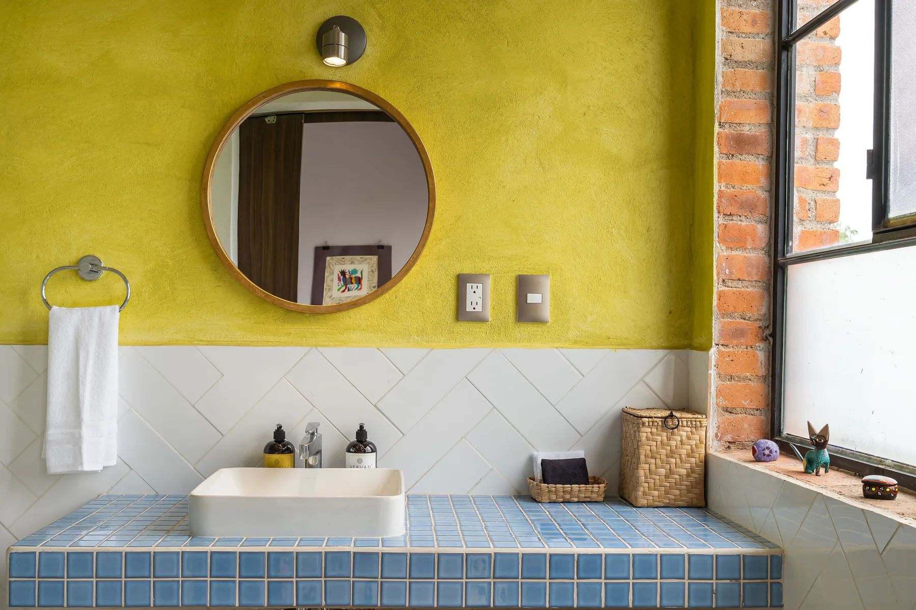 A bathroom vanity with a round mirror, tiled counter, and window.