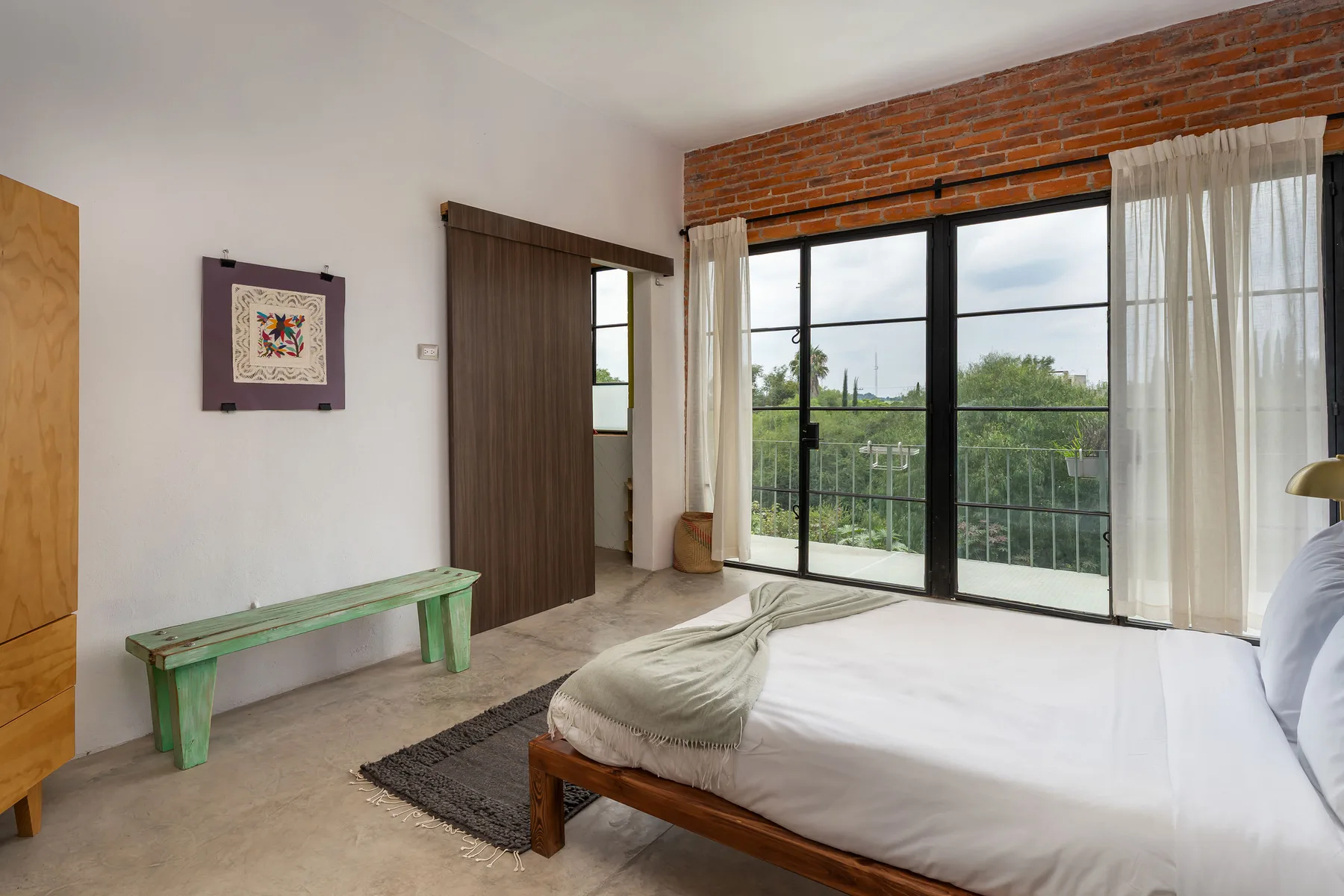 A bedroom with a bed beside large windows overlooking greenery.
