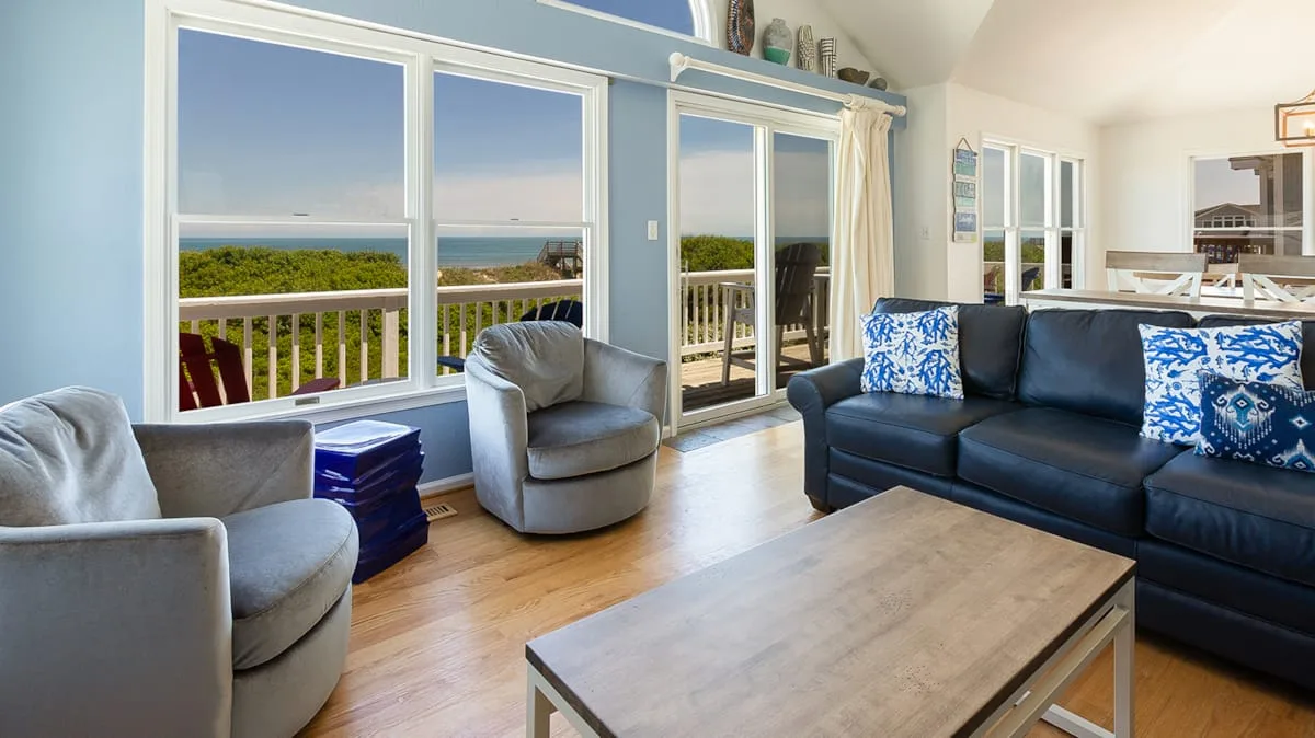 A bright living room with seating, a coffee table, and large windows overlooking greenery.