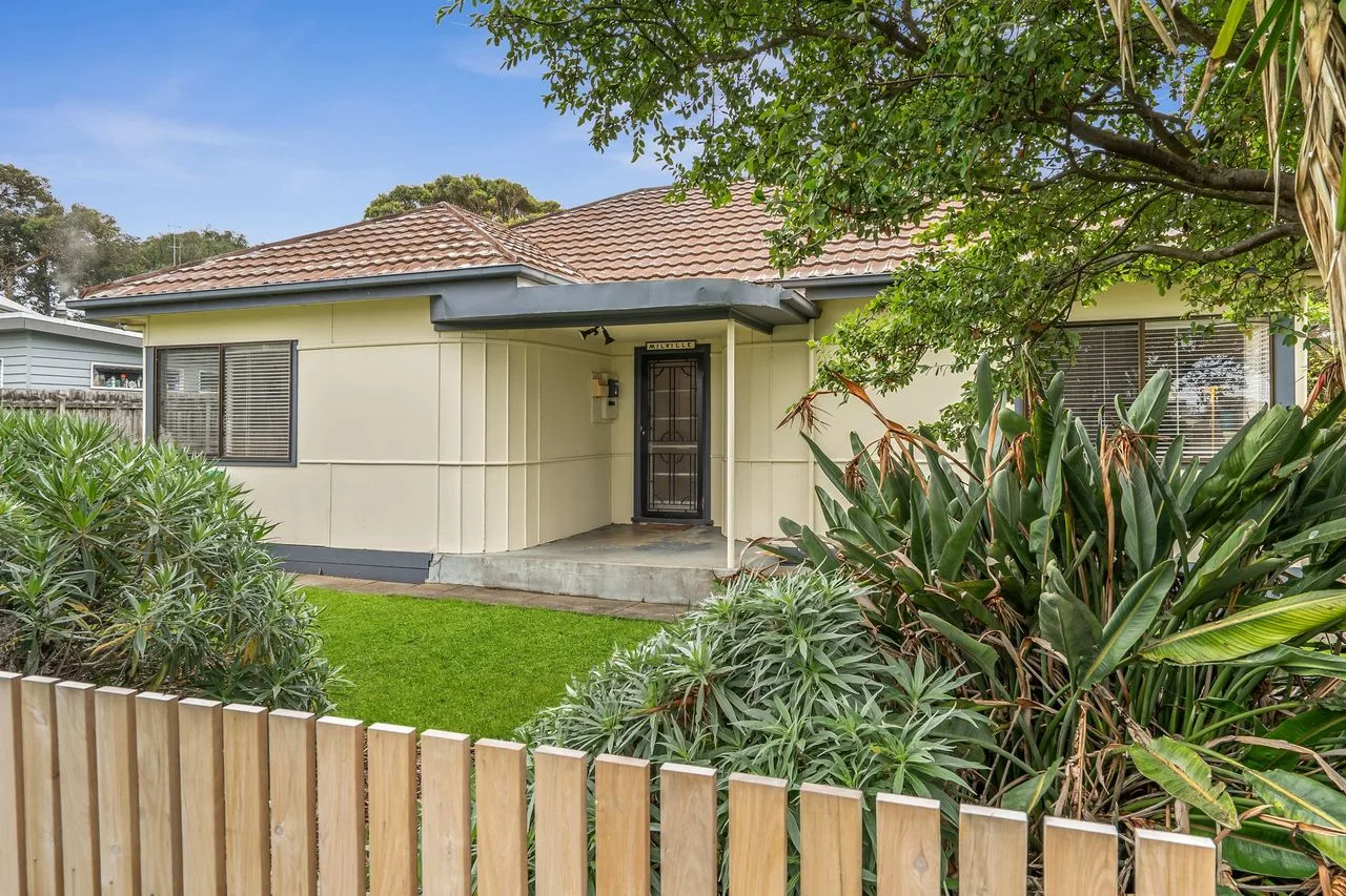 A single-story house stands behind a low wooden fence and leafy garden.