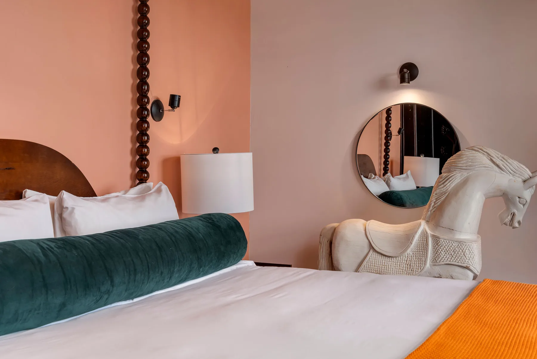 A bed with white bedding and a dark green bolster sits beside a round wall mirror.