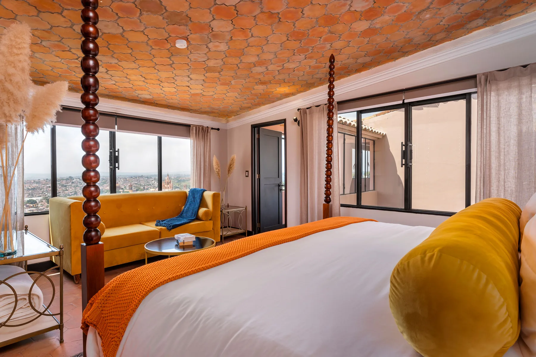 A bedroom with a large bed, mustard-colored seating, and wide windows.