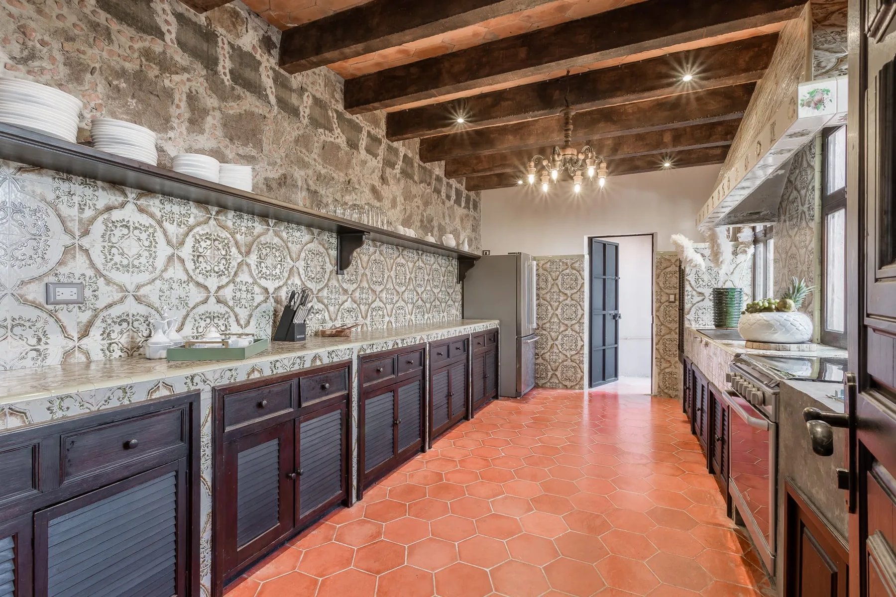 A kitchen with dark cabinets, patterned tile walls, and exposed wooden ceiling beams.