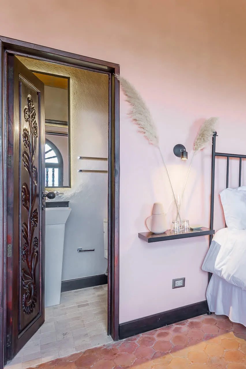 A bedroom beside an open bathroom doorway, with a wall-mounted shelf and bedside light.