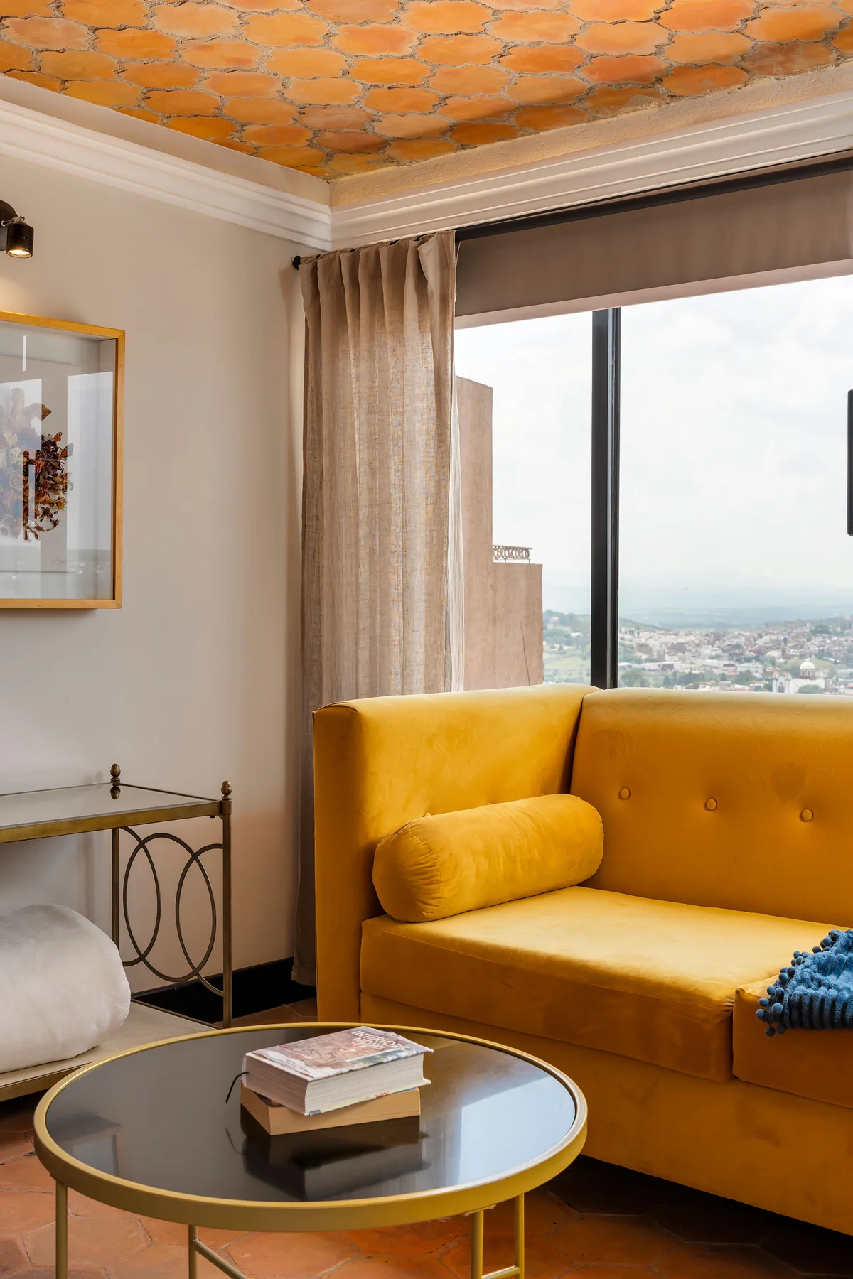 A living room with mustard-colored seating beside a large window overlooking the city.