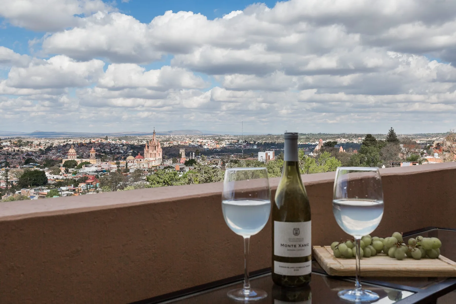 A rooftop terrace overlooks a city, with wine glasses and grapes on the table.