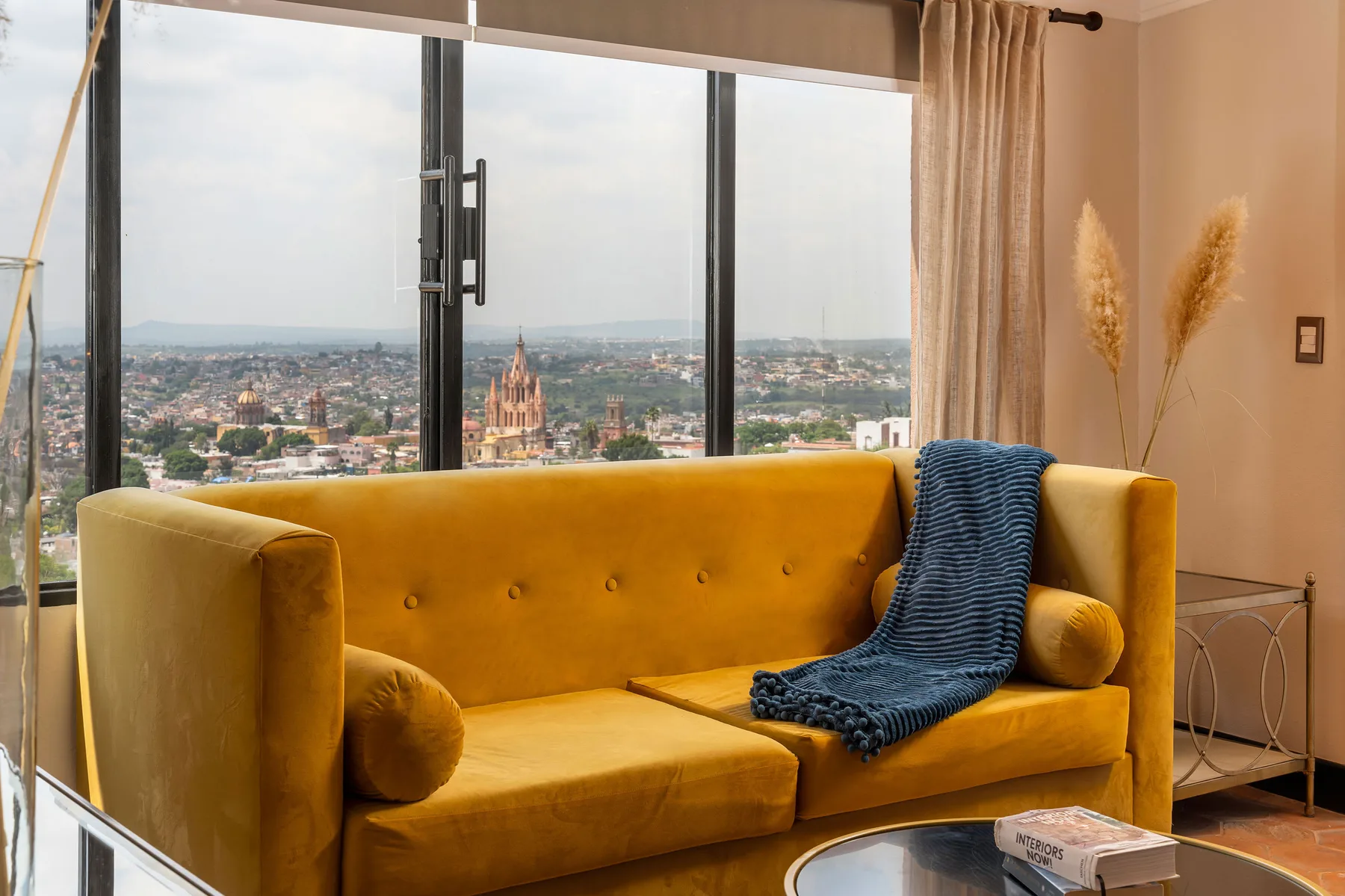 A mustard-colored sofa sits before a large window overlooking a city.