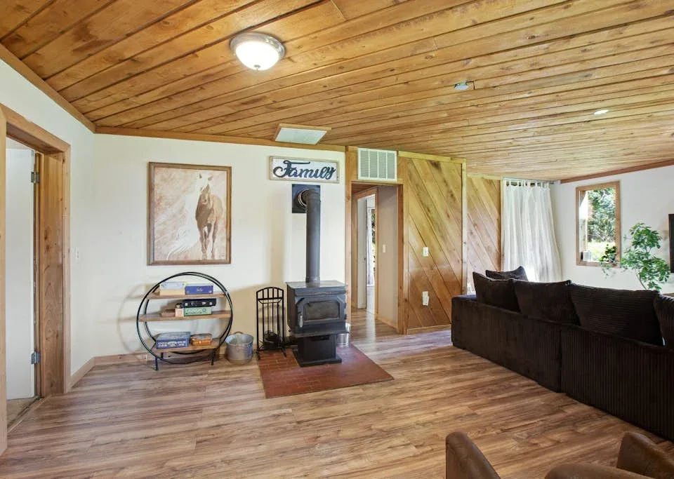 A living room with a wood stove and dark seating beneath a wood-paneled ceiling.