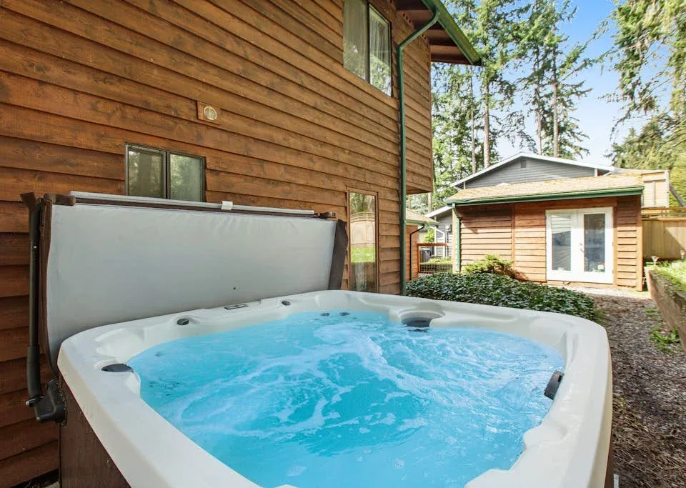 An outdoor hot tub sits beside a wooden cabin, with trees and another cabin nearby.