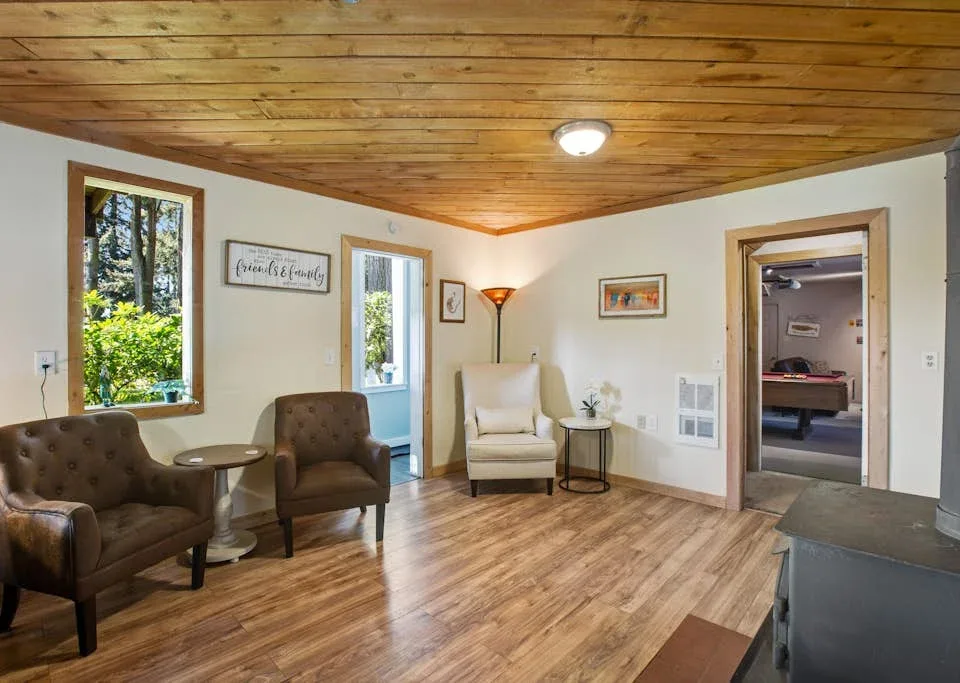 A furnished living room with seating, a wood-paneled ceiling, and windows overlooking greenery.