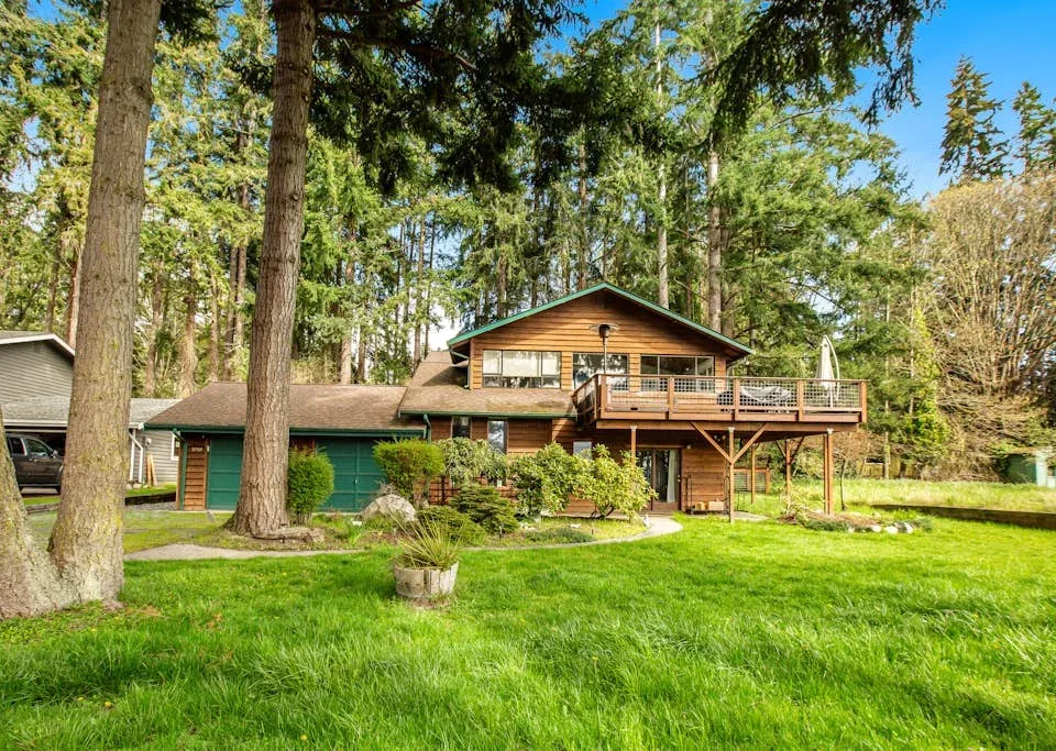 A wood-sided house with a raised deck sits in a grassy clearing among tall trees.