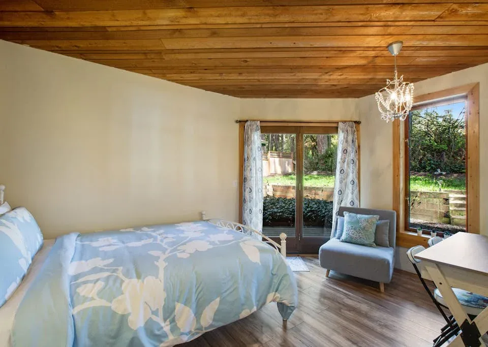 A bedroom with a bed, an armchair, and windows overlooking a garden.