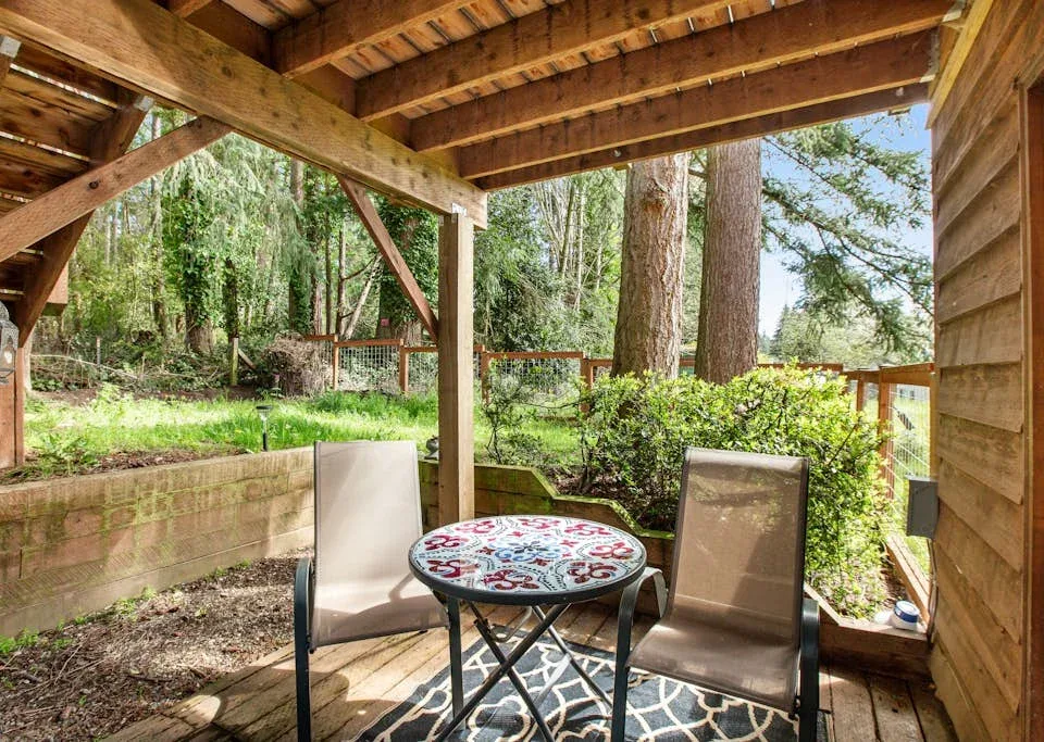 A covered wooden patio with outdoor seating overlooks a leafy wooded yard.