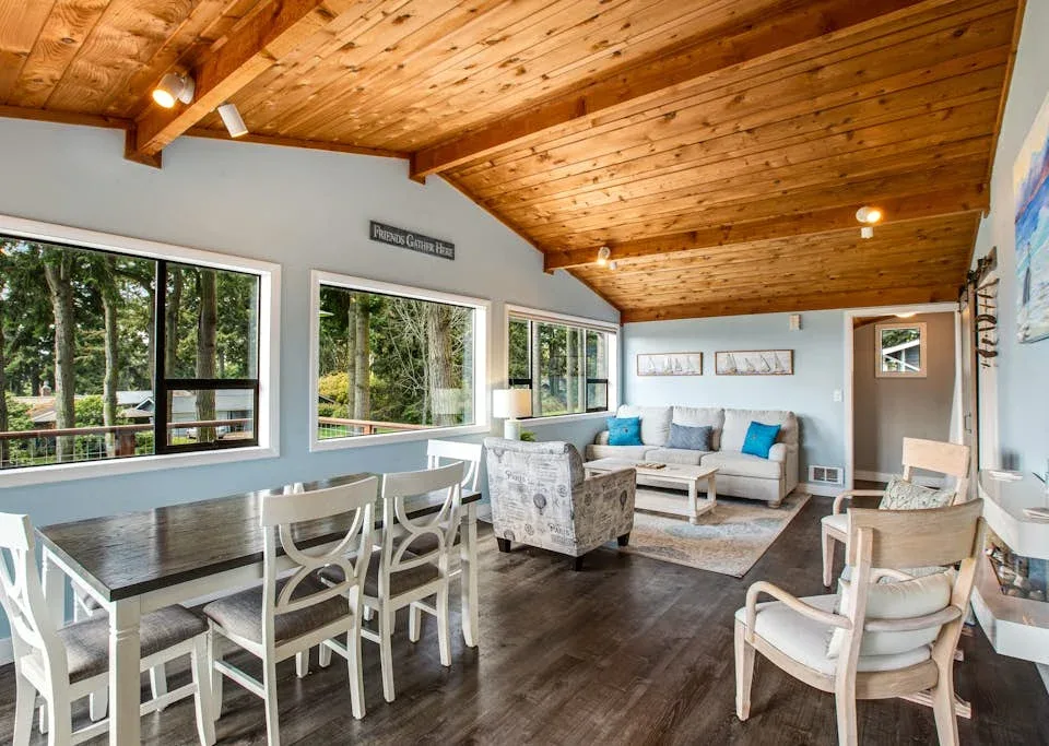 A bright living and dining area has large windows, a dining table, and seating.