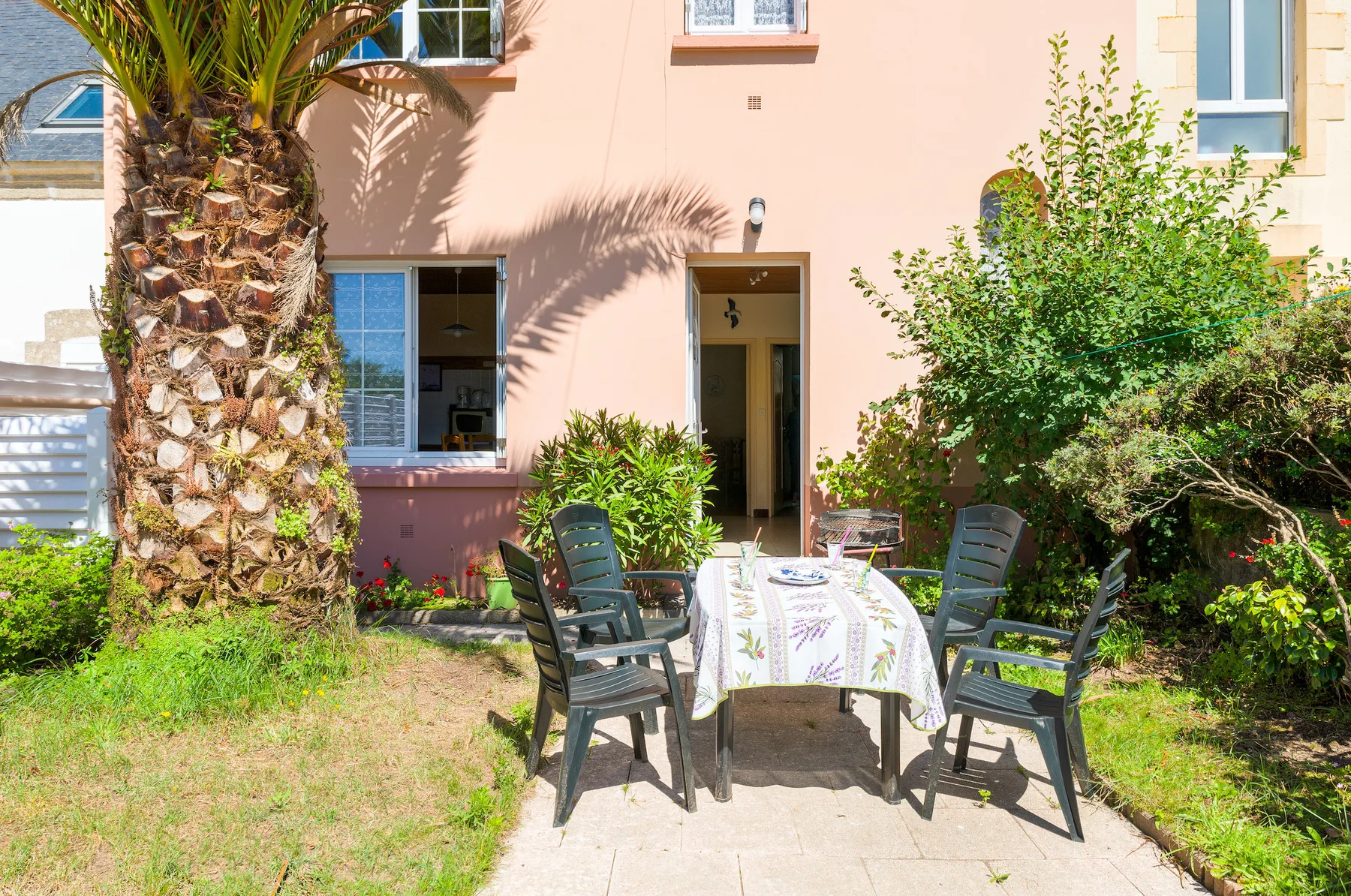An outdoor dining table with chairs sits in a garden beside a peach-colored building.