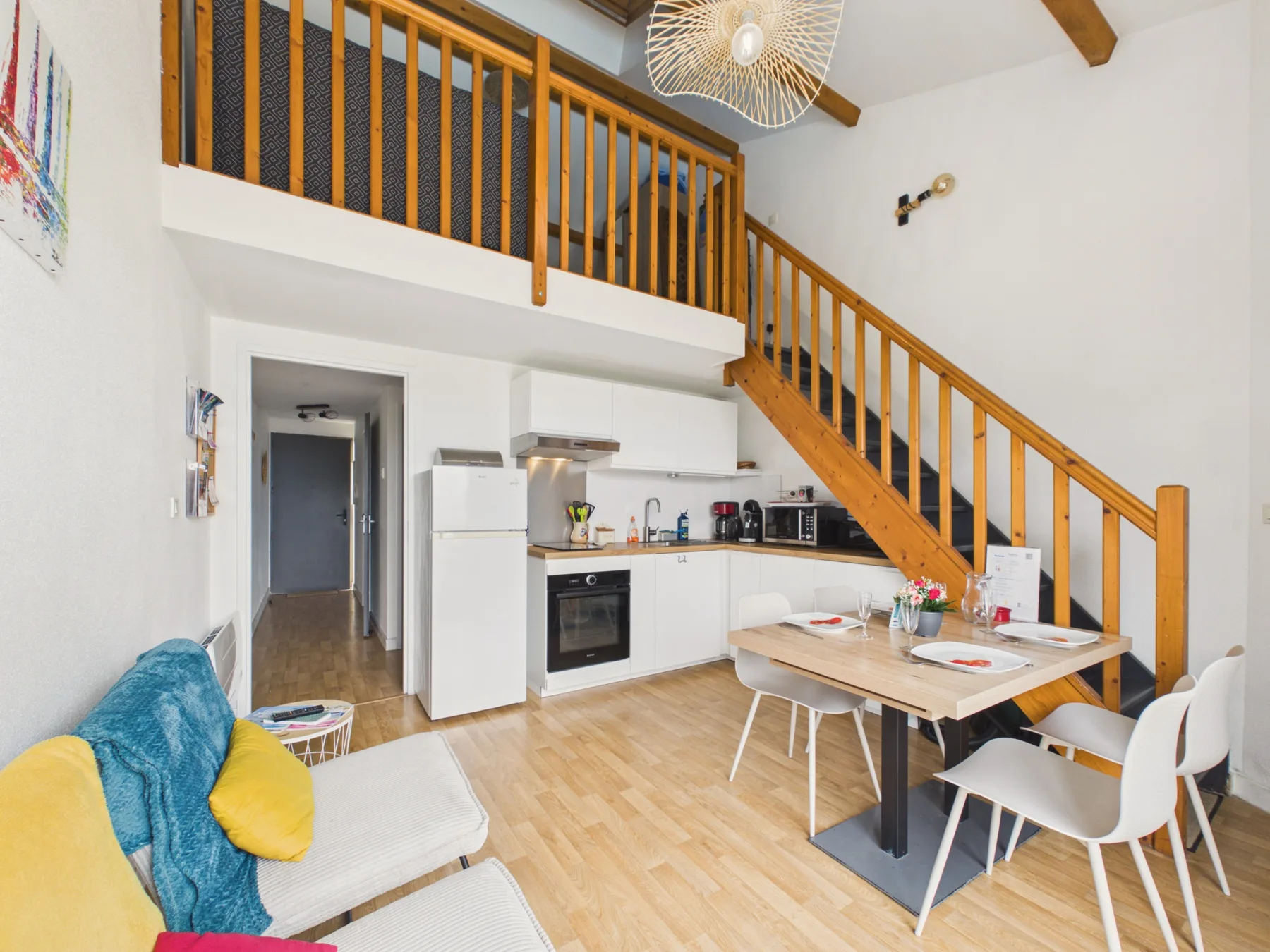 A bright open-plan living area with a kitchen, dining table, and wooden staircase.